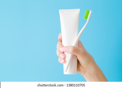 Woman's Hand Holding White Tube Of Toothpaste And Toothbrush With Green Bristles. Pastel Blue Background. Teeth Hygiene. Closeup. Empty Place For Text Or Logo.