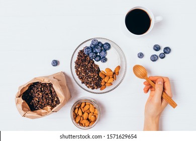 Woman's Hand Holding Wooden Spoon Near Bowl Of Granola With Yogurt, Blueberry And Almonds On White Table, Flat Lay.