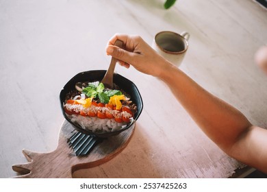 Woman's hand holding a wooden spoon above a colorful smoothie bowl with fresh fruit, seeds, and mint leaves. Rustic table and coffee cup in background, creating a serene, healthy breakfast scene - Powered by Shutterstock