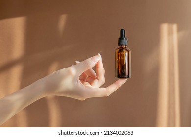 Woman's hand holds an amber glass bottle with dropper cap on brown background. Unmarked bottle with serum for face, cosmetic product. Concept of skin care and beauty.
