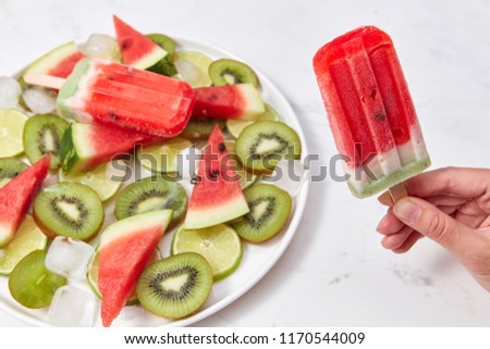 A woman's hand holds berry ice cream lolly, on a white background a plate of ice cream, pieces of kiwi, watermelon and ice cubes. Copy space for text