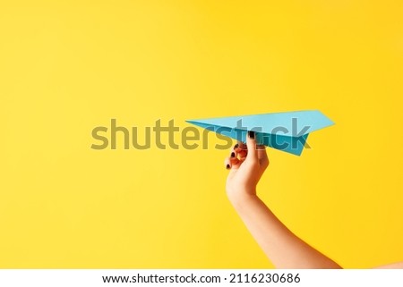 A woman's hand holds blue paper airplane on yellow background with copy space