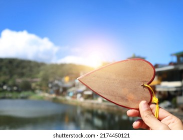 A Woman's Hand Holds A Brown Wooden Name Tag With A Red Border With A Small Heart. Raised In The Background Of The Village View And The Light That Shines In The Morning Light. - Image