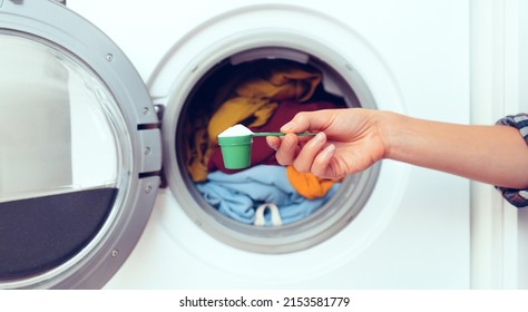 A woman's hand holds a cap with washing powder against the background of a washing machine with bright things put into it. The girl washes and wringes out things in the laundry room at home. - Shutterstock ID 2153581779