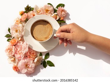 A woman's hand holds a cup of coffee. Fresh pastel peach-colored roses from the garden surround the cup. creative flower layout