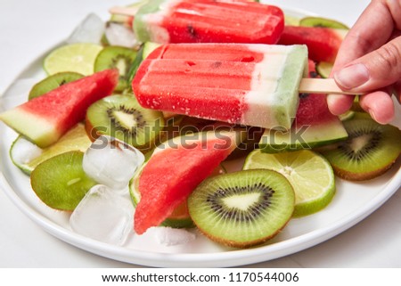 A woman's hand holds an ice cream on a stick in the form of a piece of watermelon on a background of a plate with pieces of different fruits around a white background. Close-up