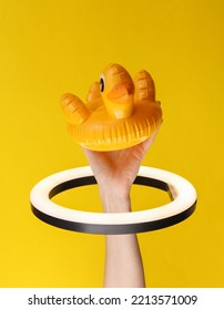 Woman's Hand Holds Inflatable Duck Through Led Ring Lamp On Yellow Background. Creative Idea