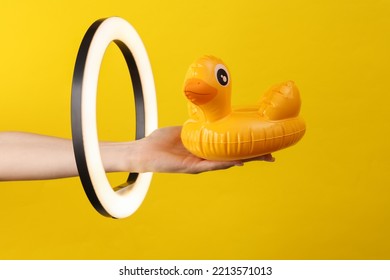 Woman's Hand Holds Inflatable Duck Through Led Ring Lamp On Yellow Background. Creative Idea