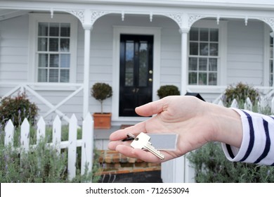 A Woman's Hand Holds A Key Against A Front Of A Traditional Villa House In Auckland, New Zealand. Buy, Sale, Real Estate, Insurance, Mortgage, Bank Loans And Housing Market Concept.