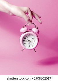 A woman's hand holds a pink alarm clock against a pastel background