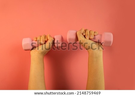 A woman's hand holds a pink dumbbell isolated on a pink background. Equipment for training at home. Fitness and activity. The concept of sport and healthy lifestyle. 