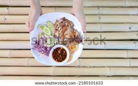 Woman's hand holds a Sliced ​​Grilled Chicken laid out in a white plate garnished with chopped cabbage, sliced ​​tomatoes, cucumbers and spicy dipping sauce with cayenne pepper served on bamboo table.