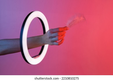Woman's hand holds soft feather through led ring lamp in red blue neon gradient light  Creative idea 