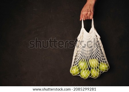 A woman's hand holds a string bag with green apples on a dark textured background