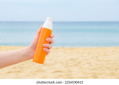 Womans hand holds a sunscreen spray in an orange bottle against the backdrop of a sandy beach and sea. The concept of applying sunscreen during a beach holiday. - Powered by Shutterstock