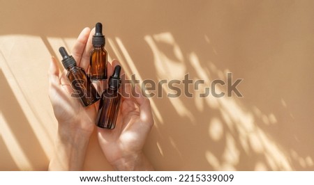 Woman's hand holds three amber glass vials with pipette on beige background in rays of sunlight. Top view of unmarked containers with essential oil, moisturizing serum. Cosmetic product for body care.