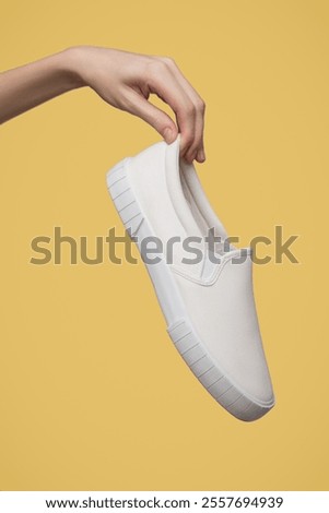 A woman's hand holds a white sneaker on yellow background.
