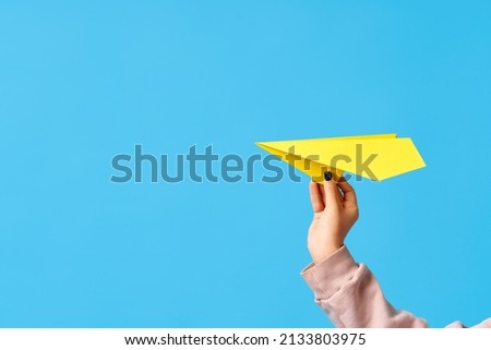 A woman's hand holds yellow paper airplane on blue background with copy space