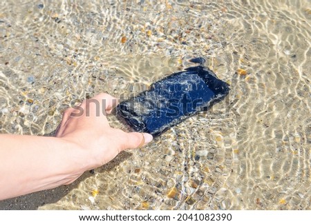 A woman's hand lifts a her smartphone from sea water on the beach. Lost and found accessories or property on vacation time, travel insurance concept image.