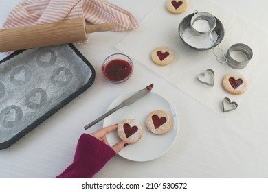 Woman's hand in lower part of frame taking a heart shaped jam filled Linzer cookie. Baking biscuits for valentines day food concept image with white background including copy space.