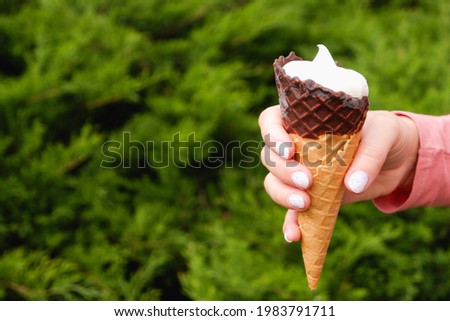 A woman's hand with a manicure holds a chocolate waffle cone of ice cream on the background of a green nature.