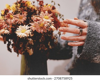 A woman's hand with a massive silver ring on a finger in a grey knitted sweater, holding a lush autumn bouquet of chrysanthemums in warm tones. The orange, yellow flowers create a cozy, autumnal scene