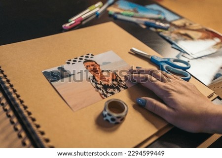Woman's hand placing a photo of a boy on the beach on top of a sheet of handmade kraft album with travel photos she is making with washi tape.