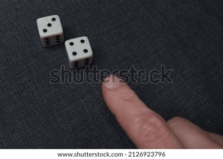 A woman's hand points to two dice from a classic board game. Numbers three and four are shown. Black and white colors predominate.