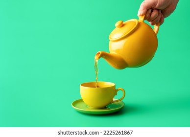 Woman's hand pour tea from yellow jug in a cup, minimalist on a green table. Hot healthy drink, mint tea in a yellow-colored cup.