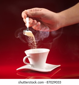 Woman's Hand Pouring Brown Sugar In White Cup Of Coffee On Red Background