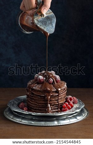 Woman's Hand Pouring Chocolate Sauce over Stack of Chocolate Pancakes with Raspberries and Red Currents