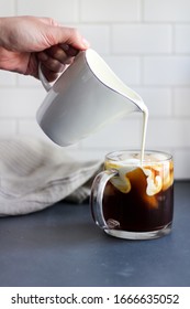 A Woman's Hand Pours Creamer Into A Glass Mug Of Iced Coffee On A Kitchen Countertop With Subway Tile Background