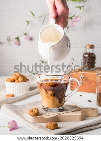
A woman's hand pours milk from a white milk jug into a glass mug with black coffee. Mixing products. Morning breakfast with dry small bagels and coffee in the kitchen on a background of white tiles. 