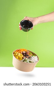A woman's hand pours soy sauce into a floating bowl of salad poke and tuna, carrot, cucumber, and pineapple with a green background. white floor - Shutterstock ID 2063103251