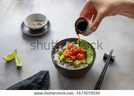 woman's hand pours soy sauce in a bowl with salmon, rice, avocado and cucumber. Food concept poke bowl. horizontal image, gray concrete background.