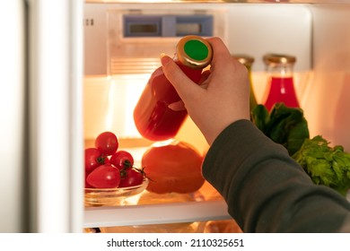 a woman's hand pulls out a glass bottle with red juice from the refrigerator
