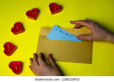 A Woman's Hand Puts A Blue Note In A Cardboard Envelope With A Handwritten Text I LOVE YOU For Her Lover On Valentine's Day. Love Letter On A Yellow Background Flatly. Red Candles In Shape Of Hearts.