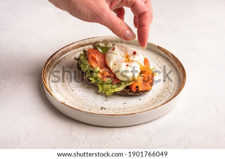 Woman's hand puts pepper on poached egg toast with avocado, salted salmon, arugula and rye bread on a ceramic plate, close up. Healthy food concept