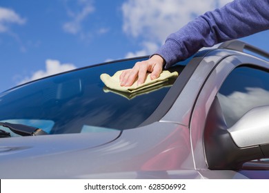 Woman's Hand With Rag Cleaning A Silver Car's Windshield On Cloudy Sky Background In Sunny Day. Early Spring Washing Or Regular Wash Up. Professional Car Wash By Hands.