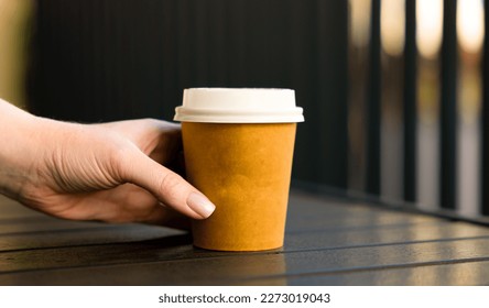 A woman's hand reaches for a paper cup of coffee with a white lid. Table on the open veranda of the cafe. - Powered by Shutterstock