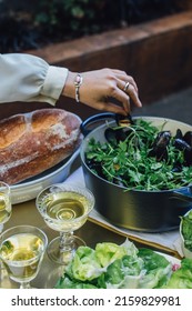 Woman's Hand Reaching Out To Take Steamed Mussel From Pot On Outdoor Dinner Table With White Wine And Bread