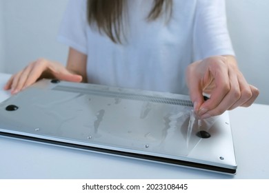 A Woman's Hand Removes The Film From A New Laptop Of Silver Color, Closeup View. Buying A Computer And Unpacking It. The Concept Of Gadgets And Computers.