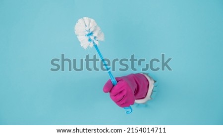 A woman's hand in a rubber glove holds a toilet brush and sticks out of a hole on a blue paper background.