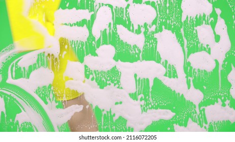 Woman's Hand In Rubber Yellow Glove Washes Window On Green Screen Chroma Key Background. Housekeeper Wipes Soap Suds From The Glass. Cleaning Windows With Cleaning Disinfectant. Close Up.