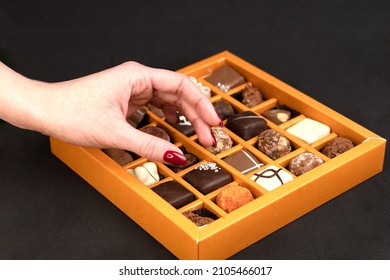 A woman's hand selects a sweet from the package. White, black, milk chocolate in an orange carton box. Tasting different types of sweets. Harmful food. Risk of obesity, diabetes and tooth decay.