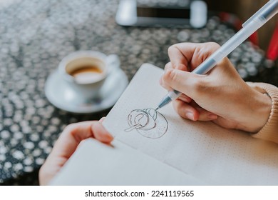 Woman's hand is sketching coffee cup on the table. Selective focus. Drawing concept. Relax concept - Shutterstock ID 2241119565