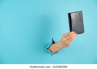 A woman's hand sticks out of a hole in a blue paper background and holds a passport in a black cover.  - Shutterstock ID 2155481981