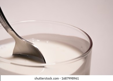 The woman's hand stirs farm yoghurt in a glass in circular motions with a spoon. Fresh soy milk in a transparent glass of white background isolated