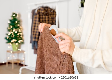 Woman's Hand Taking Knitted Sweater On Hanger On Clothes Rack In The Shop With Blurred Christmas Tree Background. Slow Fashion. Conscious Consumption. Black Friday. Winter Sale.