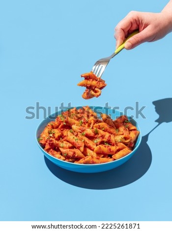 Woman's hand taking pasta with a fork from a blue bowl, minimalist on a blue table. Pasta alla arrabbiata in a blue bowl in bright light.
